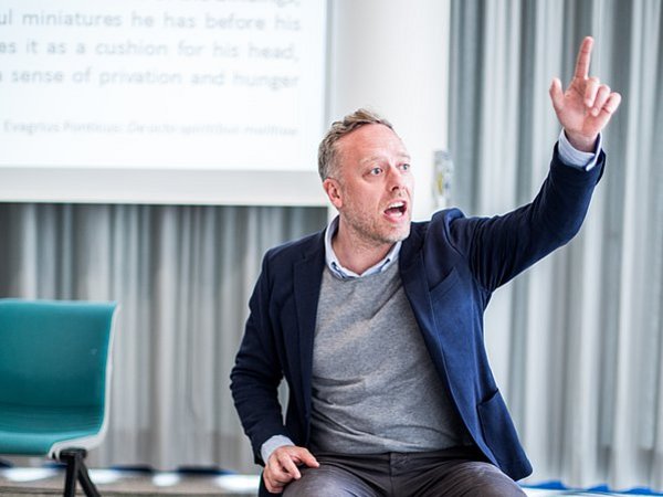 A male lecturer is sitting on a chair, talking towards the audience and gesturing wildly 