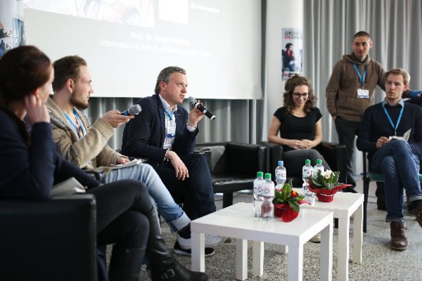 5 people are sitting in front of a large screen, discussing and speaking toward the audience